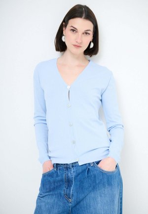 Giovane donna con capelli lisci e scuri indossa un cardigan azzurro chiaro con bottoni e jeans di denim larghi, con le mani nelle tasche, su sfondo bianco semplice.