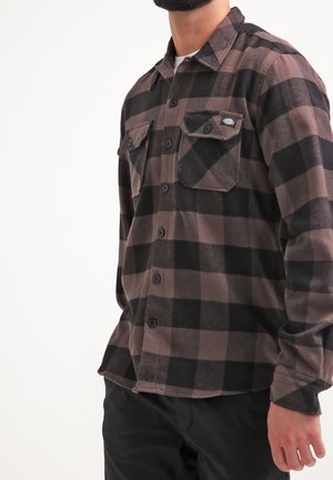 Homme portant une chemise à carreaux marron et noir avec deux poches poitrine, debout avec une main détendue le long du corps.