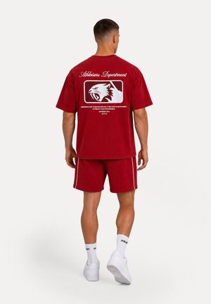 Man staat met zijn rug naar de camera en draagt een rood t-shirt en korte broek set met witte afbeelding en tekst op de achterkant, witte sokken en witte sneakers.