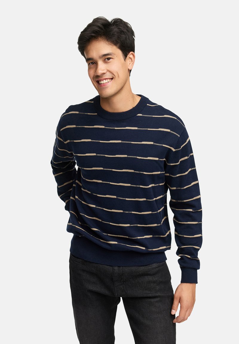 Marineblauwe trui van zacht breipatroon, met horizontale beige strepen, geribde manchetten en zoom. Het model glimlacht en draagt donkere spijkerbroek.