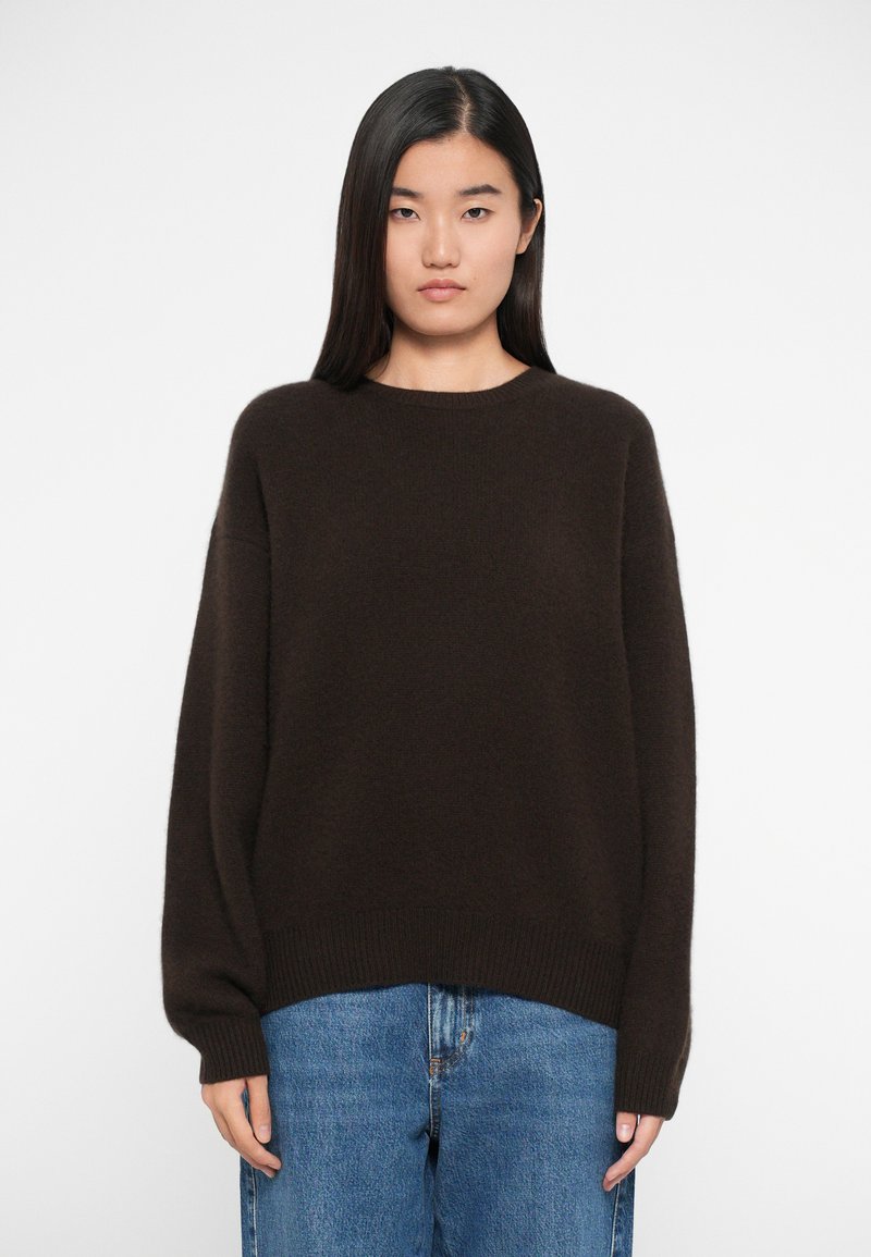 Mørkebrun strikket sweater med rund halsudskæring og sænkede skuldre. Lavet af blødt materiale med ribbede manchetter og kant. Bæres med blå jeans.