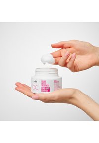 Vitt runt burk med rosa etikett, innehållande en vit krämig hårbehandling. En hand håller burken medan den andra visar en skopa av krämen.