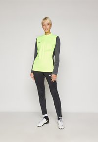 Dugih rukava atletska majica u svijetloj neonskoj žutoj boji s sivim rukavima, u kombinaciji s crnim uskim hlačama s teksturiranim detaljima i Nike logotipom.