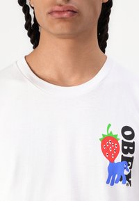 White cotton t-shirt featuring a graphic of a red strawberry with eyes and a blue elephant, along with the word "OBEY" in black.