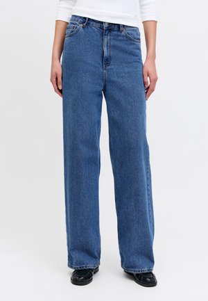 Personne portant un jean bleu taille haute coupe ample et des chaussures noires à bout carré, les mains relâchées sur les côtés et une chemise blanche à manches longues.