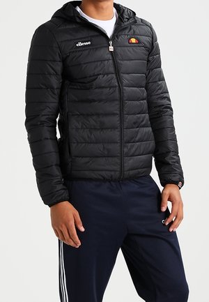 Homme portant une veste matelassée à capuche noire de la marque Ellesse et un pantalon de survêtement bleu marine avec des rayures blanches sur les côtés.