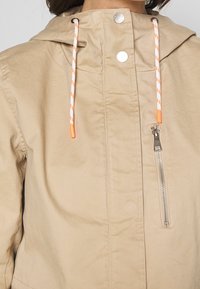 Beżowa kurtka z kapturem, z widocznym przednim zamkiem błyskawicznym, kontrastowymi pomarańczowymi sznureczkami oraz fakturowanym materiałem. Posiada zatrzaski na kołnierzu.