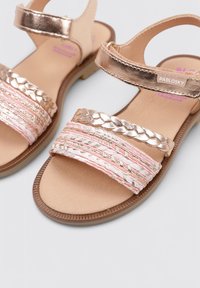 Sandalias de cuero con tiras metálicas en color rosa dorado, bandas trenzadas en tono rosa texturizado, y una suela de goma ligera. Cierre ajustable de velcro.