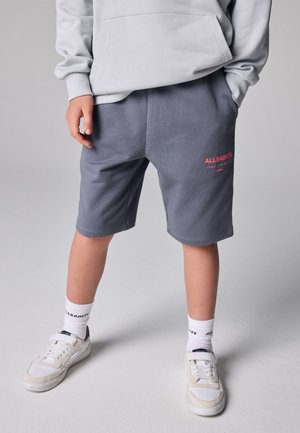 Dreng iført grå AllSaints shorts med rød logo, hvide AllSaints sokker, hvide sneakers og en lysegrå hættetrøje, stående på en grå gulv.