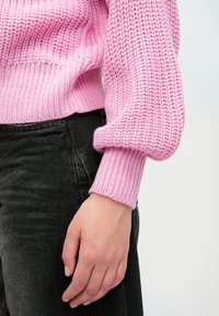 Lyserosa strikket genser med ribbekanter på hetten og ermer, med et teksturert mønster. Brukt over svarte denim.