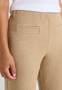 Pantalones beige texturizados con una tela tejida, que presentan un corte recto y un pequeño detalle de bolsillo trasero. Vista en primer plano de la textura de la tela.