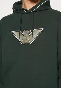 Mörkgrön huvtröja med en silver- och grå broderad logotyp. Har en dragsnodd på huven och kontrasterande piping längs halslinjen.