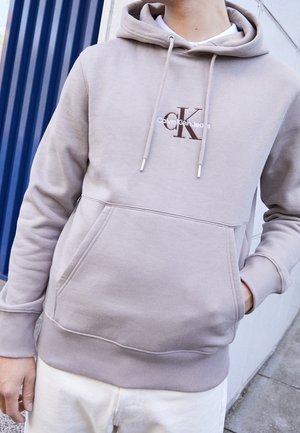 Osoba w beżowej bluzie z kapturem Calvin Klein z przednią kieszenią i białych spodniach stoi przy niebieskich i białych ścianach na zewnątrz.