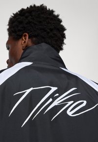 Čierna bunda s bielymi akcentmi, vybavená vysokým golierom a veľkým bielym logom "Nike" na zadnej strane. Hladká textura, atletický dizajn.