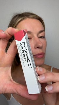 Frau hält eine MAC Locked Kiss 24-Stunden Lippenstiftbox nah an die Kamera, das Gesicht im Hintergrund leicht verschwommen.
