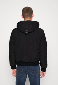 Man wearing a black hooded jacket and blue jeans seen from behind against a plain light background.