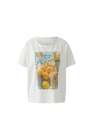 Femme aux longs cheveux foncés portant un t-shirt blanc graphique avec des fruits et du texte, une jupe midi en jean clair avec une fente devant, et des sandales beiges.