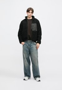 Giacca di pile nera con zip frontale, colletto alto e tasca grigia. Abbinata a jeans blu larghi e sneakers bianche. Sfondo neutro.