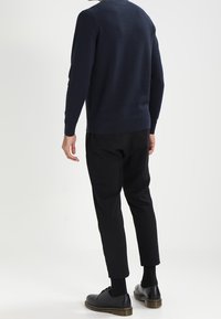 Marinblå stickad tröja med texturerat mönster, svarta smalpassade byxor och svarta snörskor. Neutral bakgrund utan synliga accessoarer.