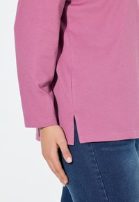 T-shirt a maniche lunghe di colore rosa chiaro con fessure laterali, realizzato in tessuto morbido. Abbinato a jeans di denim scuro. Mano appoggiata sulla coscia.