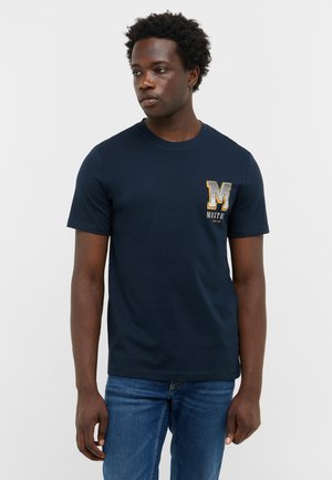 Navy blue cotton t-shirt with short sleeves, round neck, and a textured "MUST" graphic design on the chest. Worn with blue jeans.