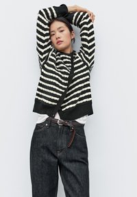 Černobíle pruhovaný svetr s ribkovanou strukturou, nošený přes bílou košili, doplněný o tmavé džíny a hnědý opasek.