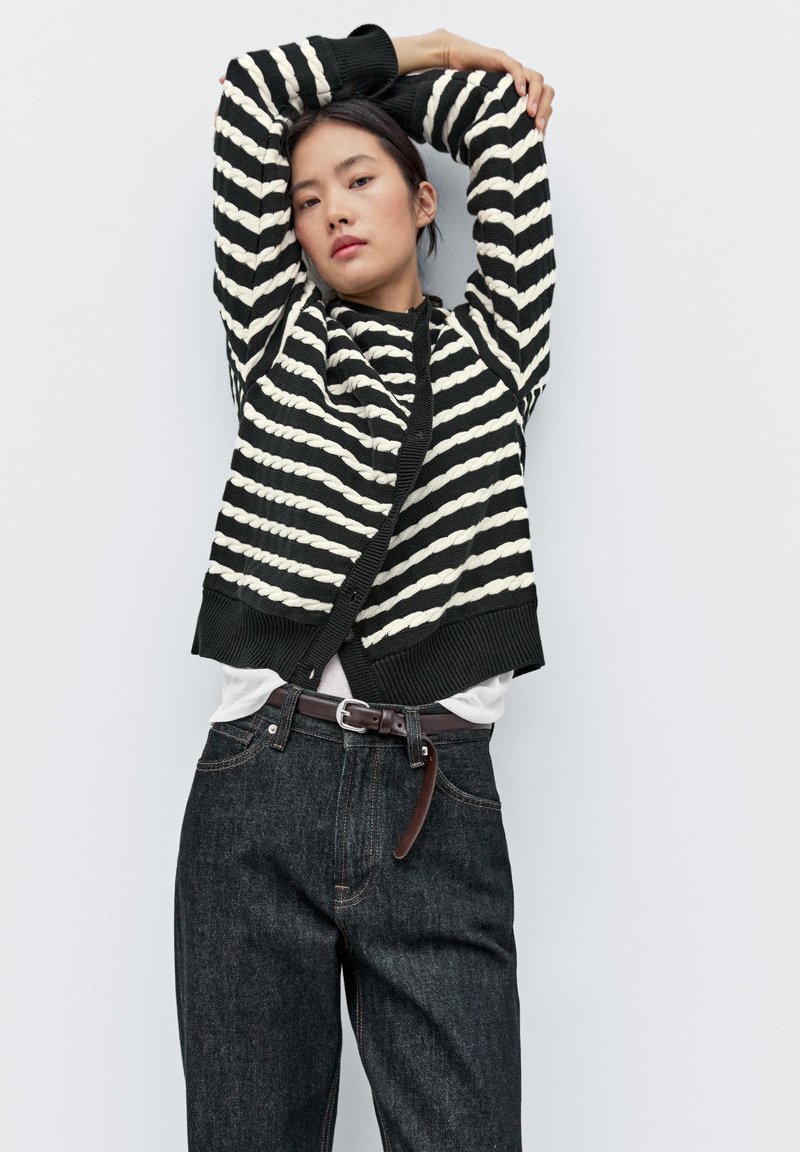 Černobíle pruhovaný svetr s ribkovanou strukturou, nošený přes bílou košili, doplněný o tmavé džíny a hnědý opasek.