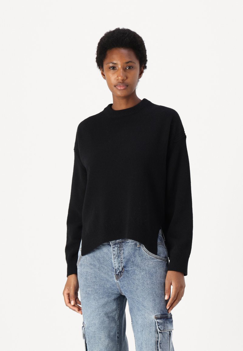 Črna pletena majica z okroglo izrezom, spuščeni rameni in rahlo skrajšanim, asimetričnim robom. Nosi se z džins hlačami s karirnimi žepi.