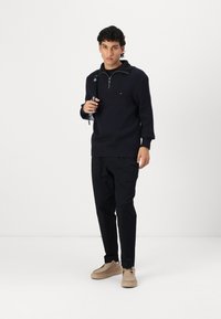 Navy gestreifter Pullover mit Reißverschlusskragen, kombiniert mit schwarzen schmal zulaufenden Hosen und beigen Schuhen. Modell hält einen blauen Taschengurt.