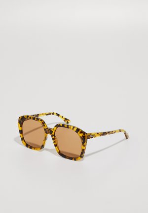 Gafas de sol carey con lentes rectangulares, un armazón grueso y lentes con un sutil degradado marrón. Presenta un logo dorado en la patilla.
