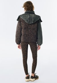 Femme portant une veste matelassée avec un imprimé léopard et des manches unies, des leggings imprimé léopard et des baskets noires, se tenant de dos.