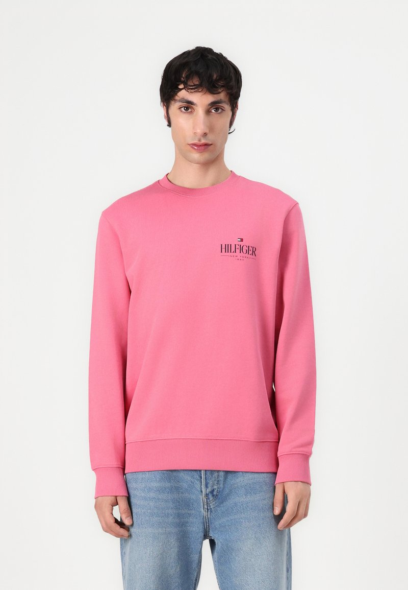 Rosa hettegenser med lange ermer, rund hals, ribbede mansjetter og kant, med en liten svart logo på brystet, brukt sammen med lyseblå jeans.