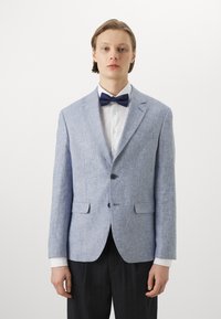 Gaiši zils bleizeris ar strukturētu audumu, vienas pogas dizains, divas priekšējās kabatas un tumši zils tauriņš. Zem tā redzams balts krekls.