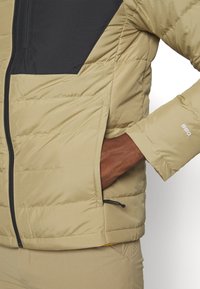 Veste isolante beige avec accents noirs, dotée d'une fermeture éclair et d'une poche latérale. Tissu texturé avec coutures matelassées visibles.