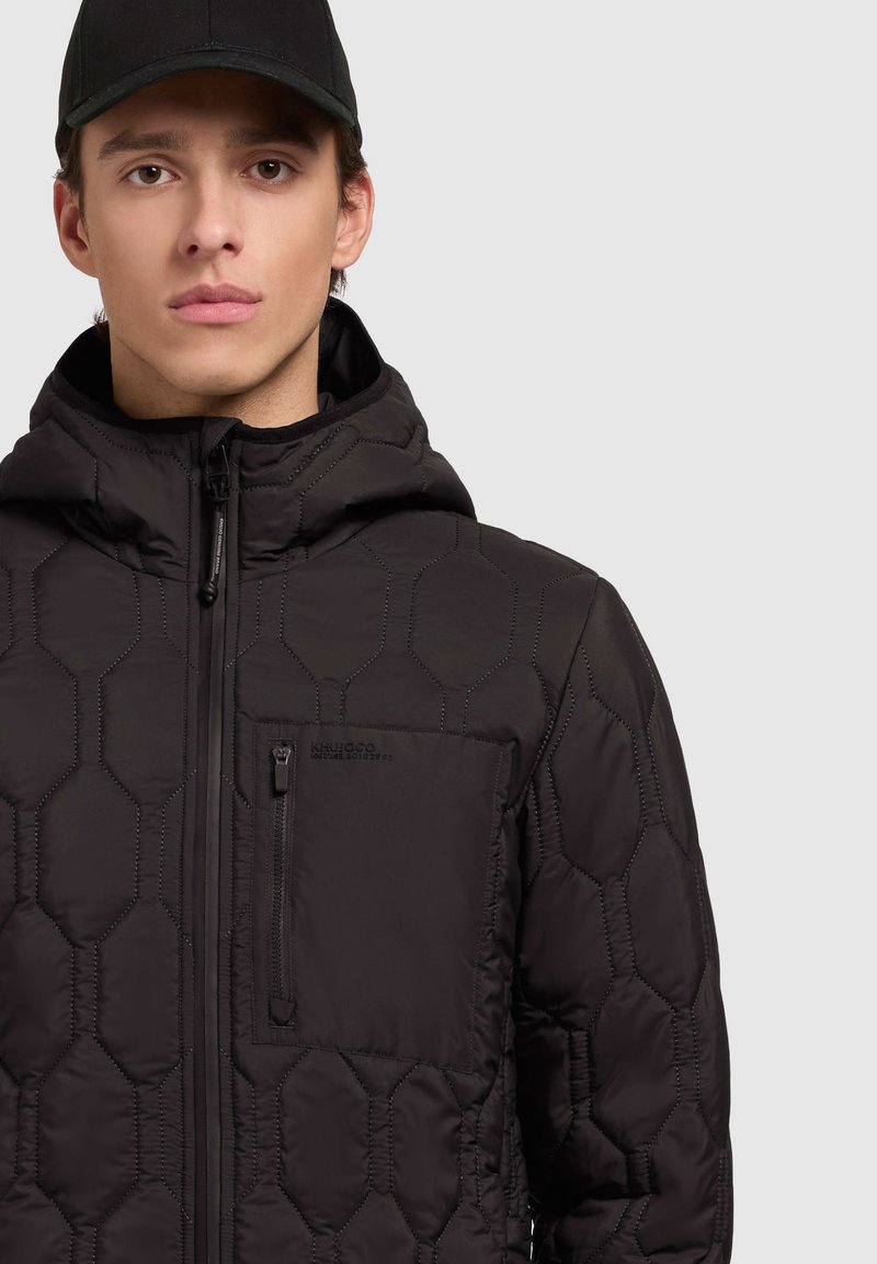 Veste matelassée noire avec un motif hexagonal, fermeture éclair à l'avant, capuche réglable et poche poitrine zippée. Le tissu semble texturé et léger.