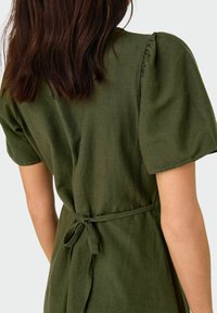 Olivgrön klänning gjord av ett texturerat tyg, med korta puffärmar och ett band i midjan, har en figurnära bodice och en flödande kjol.