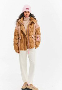 Kobieta w beżowej pikowanej kurtce z różowymi akcentami, jasnobeżowych spodniach, beżowych butach turystycznych i różowej czapce, stojąca na białym tle.