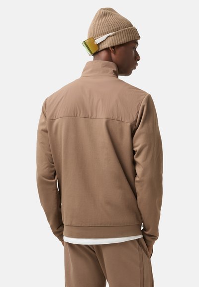 Brown athletic jacket with a smooth, flexible texture, high collar, and matching tan pants. Accessories include a beige knit hat with a colored accent.