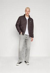 Svart dunjakke med horisontale sømmer, glidelåslukking og liten blå logo på brystet, kombinert med lysegrå jeans og svarte joggesko.