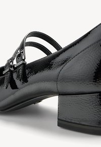 Chaussures basses à talon carré en cuir verni noir, avec doubles brides à boucles, bout arrondi et finition texturée, vue angle mettant en valeur le talon.
