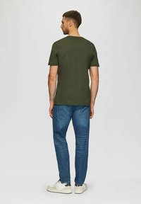Mann steht mit dem Rücken zur Kamera, trägt ein olivgrünes T-Shirt, blaue Jeans und weiße Sneakers vor einem schlichten hellen Hintergrund.