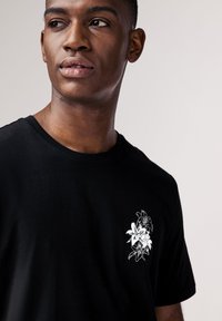 Schwarzes Baumwoll-T-Shirt mit einem weißen Blumenprint auf der linken Brust. Klassischer Rundhalsausschnitt, kurze Ärmel und angenehme Textur.