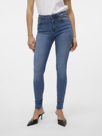 Ženska v modrih skinny jeans hlačah, beli majici in črnih špičastih visokih petah stoji ob nevtralni ozadju.