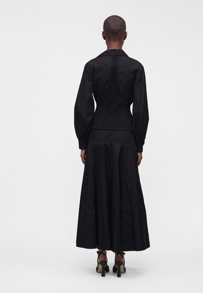 Personne aux cheveux courts portant une veste noire ajustée et une longue jupe noire avec des sandales à talons, debout de dos sur un fond blanc.
