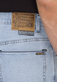 Jean de mezclilla azul claro con un parche de cuero, que presenta diseños gráficos y texto. Incluye una pequeña etiqueta negra "VOLCOM" en el bolsillo.