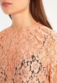 Perzikkleurige kanttop met bloemmotieven, een ronde halslijn en zachte plooien rond de kraag, gelaagd over een donkerder stof.