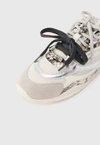 Αθλητικό παπούτσι με μπεζ πλέγμα στο επάνω μέρος, ασημένιες λεπτομέρειες, κορδόνια σε μαύρο και μπεζ, και πλευρικό πάνελ με μοτίβο ερπετού. Διακριτικό λογότυπο στα κορδόνια.