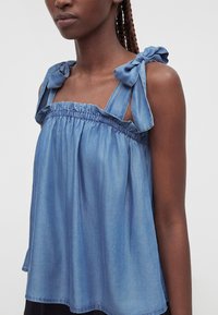 Femme portant un haut bleu clair sans manches avec un col froncé et des bretelles nouées aux épaules, cheveux tressés visibles d'un côté.