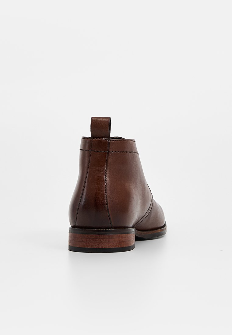 Botas de tobillo de cuero marrón con una textura suave, una tira en la parte posterior y un tacón de madera apilada. Detalles de costura mínimos.
