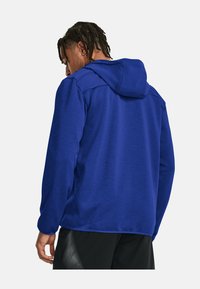 Blauer Kapuzenpullover mit strukturiertem Finish, ausgestattet mit einer Kordelzugkapuze und einem entspannten Schnitt. Sichtbare schwarze Shorts mit einem atmungsaktiven Design.
